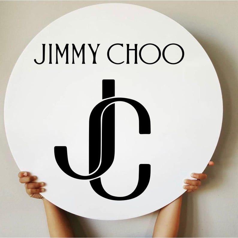 May include: A white, round sign featuring the black text "JIMMY CHOO" above a stylized "JC" logo. The logo is a black, interlocking design of the letters J and C. The sign is held up by two hands.
