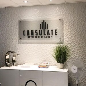 May include: A modern office reception area featuring a white textured wall and a sign that reads "CONSULATE DEVELOPMENT GROUP." A white cabinet holds decorative items and a potted plant. A fan is visible.