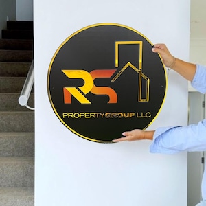 Może przedstawiać: Okrągły znak w kolorze czarnym i złotym z literami "RS" w gradiencie ż&oacute;łtym i pomarańczowym, stylizowanym ikoną domu i tekstem "PROPERTY GROUP LLC".