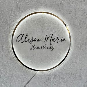 May include: A round, illuminated sign with the text "Alison Marie Hair & Beauty" in elegant script. The sign features a gold-colored outer ring and a bright, white inner glow, mounted on a textured, light-colored wall.