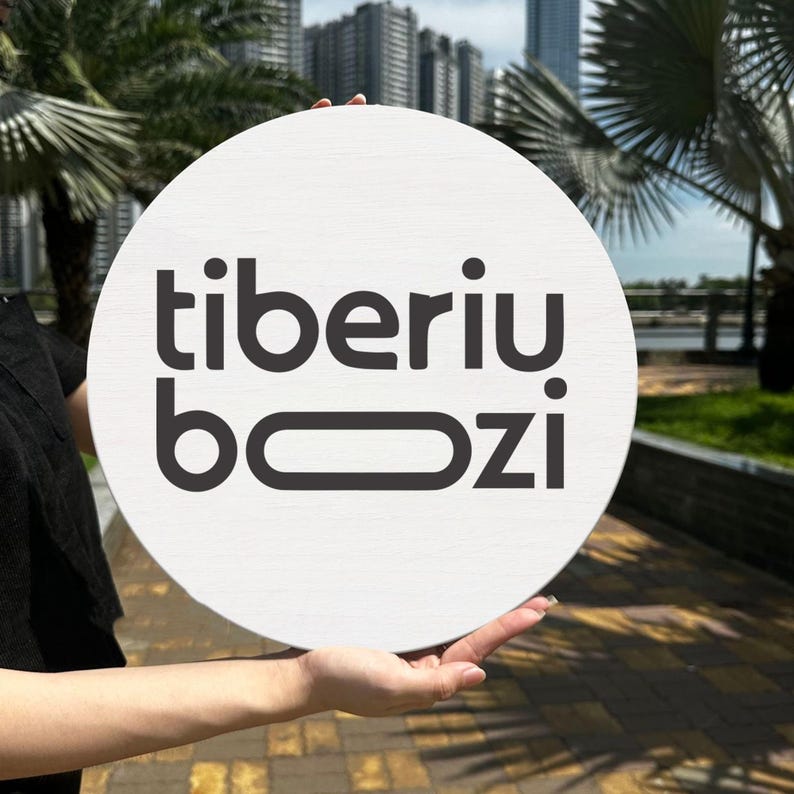 May include: A round, white sign with the black text "tiberiu bozi" in a bold, sans-serif font. The sign is held up against a blurred outdoor background with palm trees and buildings.