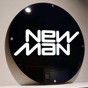 May include: A round, black sign with the white text "NEW MAN". The sign is mounted with four silver screws. The sign is glossy and reflects the light.