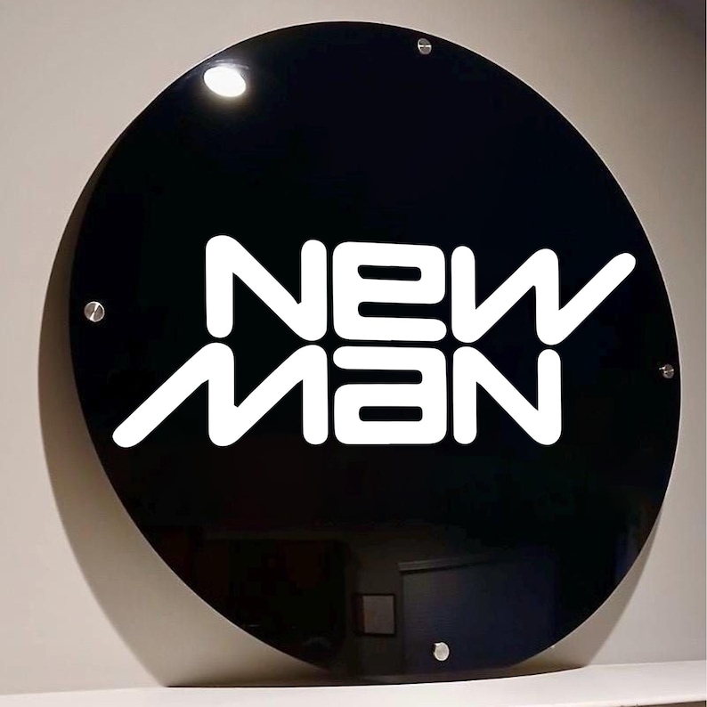May include: A round, black sign with the white text "NEW MAN". The sign is mounted with four silver screws. The sign is glossy and reflects the light.