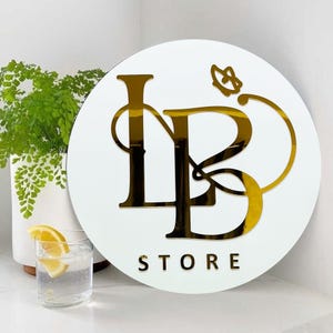 Może przedstawiać: Okrągły znak w kolorze białym z logo w kolorze złotym, przedstawiającym splecione litery "LB" z ozdobnym wzorem kwiatowym. Pod logo znajduje się napis "STORE" w kolorze złotym.