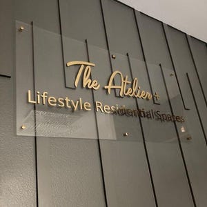 以下が含まれることがあります： 「The Atelier + Lifestyle Residential Spaces」と金色の文字で書かれた透明なアクリルサイン。 金色の金具が付いた灰色のパネル壁に取り付けられています。