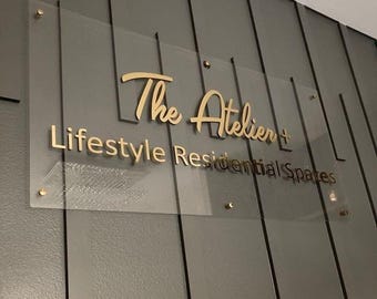 Może przedstawiać: Przezroczysty akrylowy szyld ze złotym napisem "The Atelier + Lifestyle Residential Spaces". Szyld jest zamontowany na szarej ścianie z panelami ze złotym osprzętem.