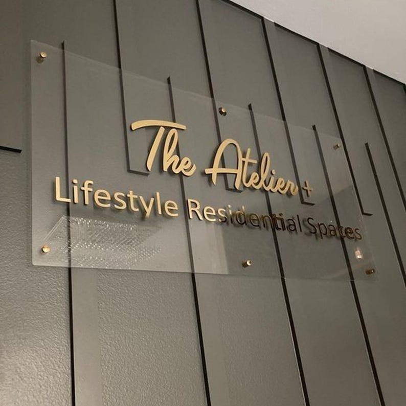 May include: A clear acrylic sign with gold lettering that reads "The Atelier + Lifestyle Residential Spaces." The sign is mounted on a gray paneled wall with gold-colored hardware.