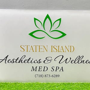 May include: A rectangular sign with a white background features the words "STATEN ISLAND Aesthetics & Wellness MED SPA" in gold and black script. A green lotus flower logo is at the top, and a phone number is at the bottom.