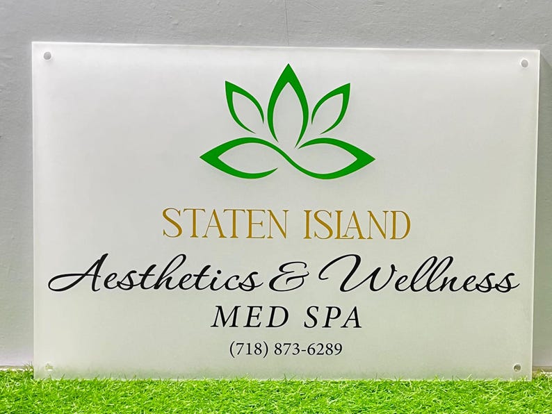May include: A rectangular sign with a white background features the words "STATEN ISLAND Aesthetics & Wellness MED SPA" in gold and black script. A green lotus flower logo is at the top, and a phone number is at the bottom.