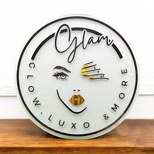Może przedstawiać: Okrągły biały znak z czarnym napisem "Glam Glow, Luxo & More" ze stylizowaną ilustracją oka i ust kobiety z złotym lakierem do paznokci i ustami.