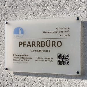 May include: A rectangular sign with the text "PFARRB&Uuml;RO" in large, bold letters. The sign also includes the address "Danhauserplatz 2", opening hours, a QR code, and the logo of the "Pfarreiengemeinschaft Aichach". The sign is mounted on a textured wall.