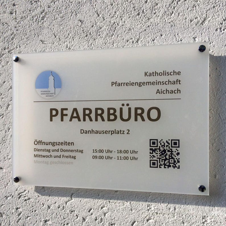 May include: A rectangular sign with the text "PFARRB&Uuml;RO" in large, bold letters. The sign also includes the address "Danhauserplatz 2", opening hours, a QR code, and the logo of the "Pfarreiengemeinschaft Aichach". The sign is mounted on a textured wall.