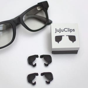May include: Black eyeglasses with a square frame are positioned above a small white box with the text "JujuClips". Four black clips are below the box. The clips are designed to attach to the eyeglasses.