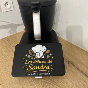 May include: A black kitchen appliance with a silver handle and a rectangular black mat in front. The mat features a white chef hat, a whisk, a wooden spoon, and the text "Les délices de Sandra" with "conseillère thermomix" below.