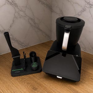 May include: A black Thermomix kitchen appliance with a digital display, a black utensil holder, and a small succulent plant in a pot. The appliance has a silver handle and is on a wooden surface.