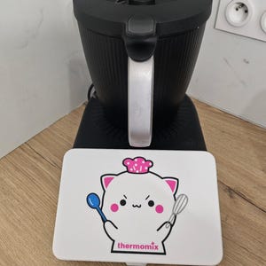 Puede incluir: Un electrodoméstico de cocina negro con mango plateado y una cubierta blanca rectangular que presenta un gato de dibujos animados con un gorro de chef rosa, sosteniendo una cuchara azul y un batidor, con la palabra thermomix impresa en rosa.
