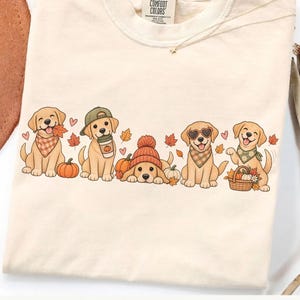 Puede incluir: Una camiseta de color crema con un gráfico de cinco cachorros golden retriever con ropa de otoño. Los cachorros aparecen con calabazas, hojas de otoño, una taza de café, un gorro de punto, gafas de sol en forma de corazón y una cesta.