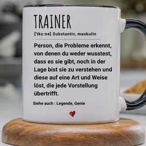 Pode incluir: Caneca de cerâmica branca com borda e alça pretas, com a palavra "TRAINER" em letras pretas em negrito. A caneca tem uma definição em alemão, com um pequeno símbolo de coração vermelho na parte inferior. A caneca está em um descanso de madeira.