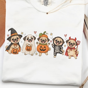 Pode incluir: Uma t-shirt branca com um gráfico de cinco cães pug vestidos com fantasias de Halloween, incluindo uma bruxa, um fantasma, uma abóbora, um esqueleto e um diabo. O design inclui pequenas abóboras cor de laranja, corações vermelhos e estrelas amarelas.