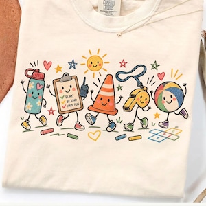 May include: A cream-colored t-shirt featuring a colorful graphic of anthropomorphic school and play items including a water bottle, clipboard with text PLAY BE KIND HAVE FUN, walkie-talkie, traffic cone, whistle, and beach ball with stars.