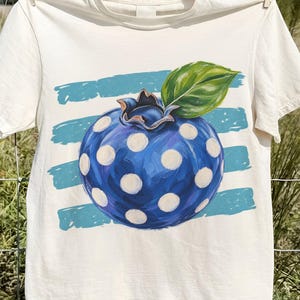 May include: A cream-colored short-sleeve t-shirt featuring a large, hand-painted style graphic of a blue blueberry with white polka dots and a single green leaf. The blueberry is centered over a background of four horizontal light blue brushstrokes.