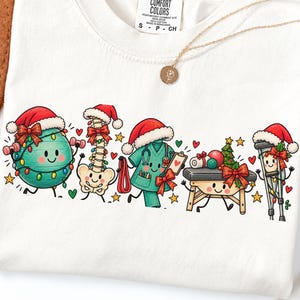 Op de afbeelding: Een wit t-shirt met een afbeelding van vijf antropomorfe medische voorwerpen met kerstmutsen: een groene fitnessbal, een ruggengraat, een groenblauw medisch shirt, een massagetafel en krukken, allemaal versierd met kerstverlichting en strikken.