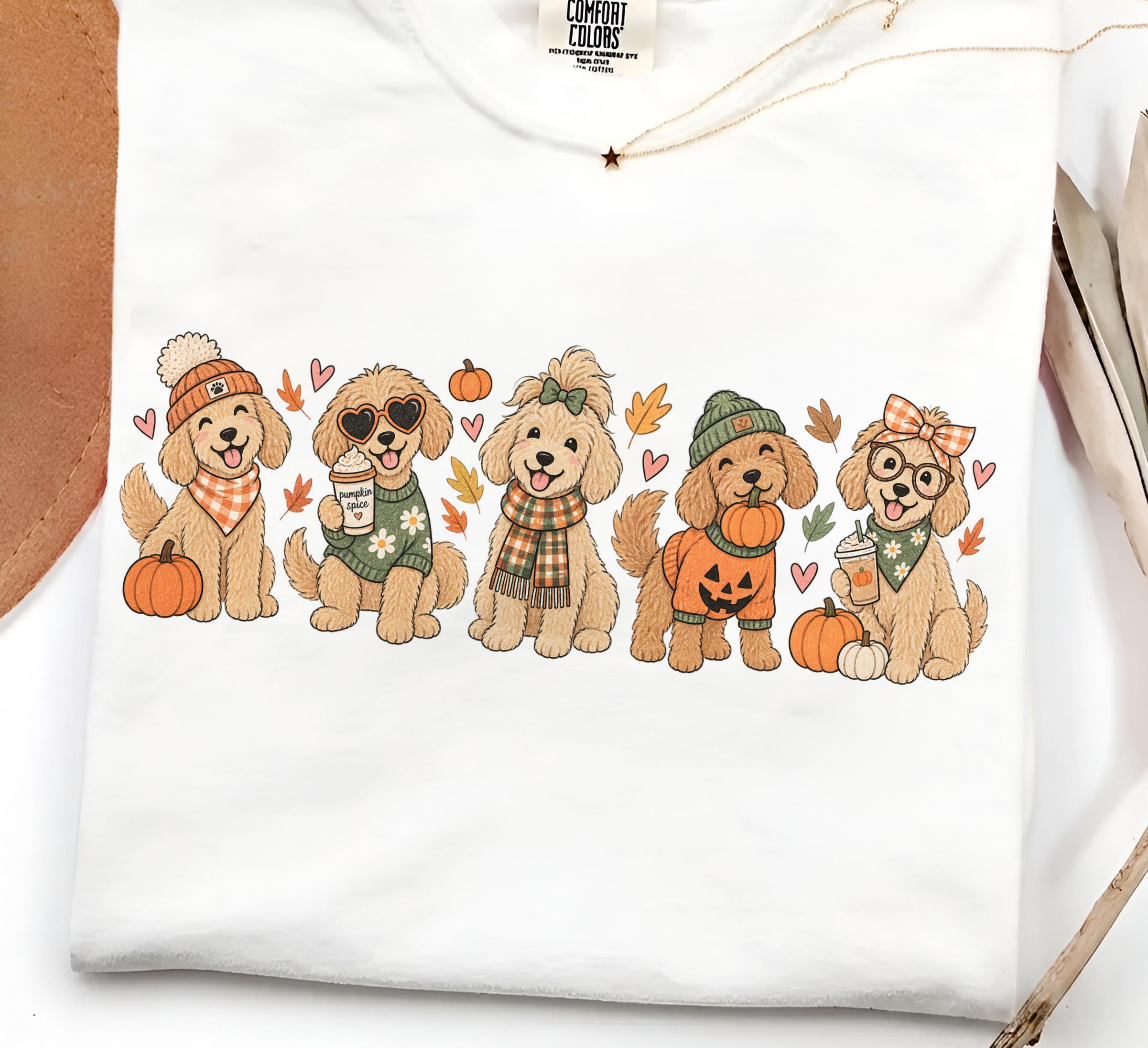 May include: A white t-shirt featuring a graphic print of five golden-colored dogs dressed in autumn-themed clothing and accessories. The dogs are surrounded by small pumpkins, fall leaves, and hearts. The shirt has a Comfort Colors brand label.