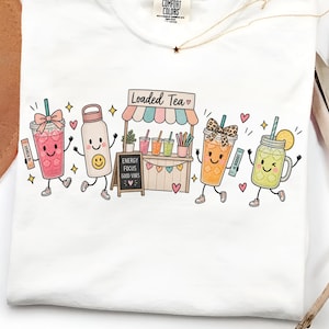 Puede incluir: Una camiseta blanca con un estampado gráfico de bebidas antropomórficas y un puesto con la etiqueta Loaded Tea. El diseño incluye una bebida rosa con un lazo, una botella de agua blanca con una cara sonriente, una bebida naranja con un lazo de leopardo y una bebida verde.
