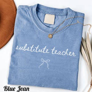 May include: A blue jean colored t-shirt featuring the white text substitute teacher written in a cursive font above a simple white line drawing of a bow. A gold chain necklace with a small round pendant rests on the collar area.