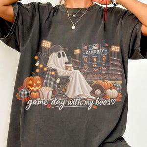 May include: A charcoal gray t-shirt featuring a graphic of a ghost wearing a baseball cap and holding a coffee cup while sitting in a stadium chair. The print includes a scoreboard reading GAME DAY, pumpkins, popcorn, and the text game day with my boos.