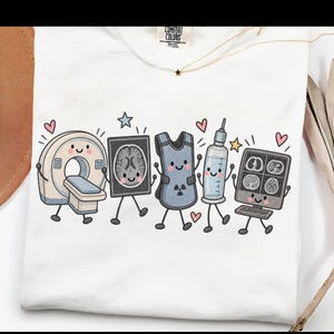 May include: A white t-shirt featuring a graphic print of five anthropomorphic medical items: a CT scanner, a brain scan, a lead apron, a syringe, and a computer monitor displaying medical images, all with smiling faces, stars, and pink hearts.