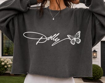 May include: A charcoal gray long-sleeve crewneck sweatshirt with a raw-edge hem. The front features white screen-printed text that reads Dolly in a cursive script, followed by a detailed graphic of a butterfly with open wings.