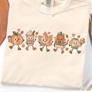 Pode incluir: Uma t-shirt de cor creme com uma impressão gráfica de cinco personagens antropomórficos com temática de outono: uma abóbora, uma chávena de café, uma folha de ácer, uma pilha de livros com uma abóbora no topo e uma camisola, com corações.
