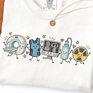 May include: A white t-shirt featuring a graphic print of five anthropomorphic medical imaging items: a CT scanner, a lead apron, a monitor displaying scans, an injector, and a radiation symbol, all decorated with small colourful hearts and stars.