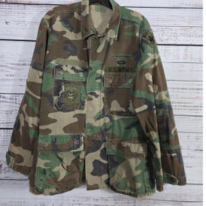 May include: Camouflage pattern jacket with multiple pockets and the text "U.S. ARMY". The jacket features a green, brown, and tan color scheme. Patches are visible on the chest and sleeve. The jacket has a button-up front and a collar.