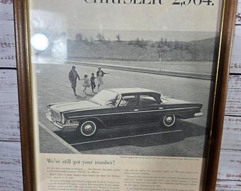 May include: Framed vintage Chrysler advertisement. The black and white image features a classic car, a family, and text that reads "CHRYSLER $2,964." The frame is a dark wood color, adding a touch of nostalgia to the print.