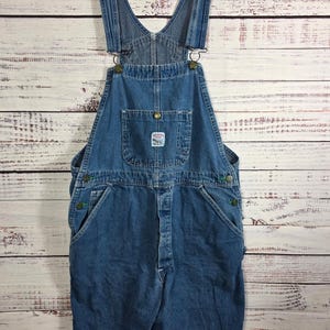 May include: Blue denim overalls featuring adjustable shoulder straps with metal buckles, a front bib pocket with a small rectangular brand patch, side button closures, and two deep hip pockets. The garment has a classic straight-leg cut.