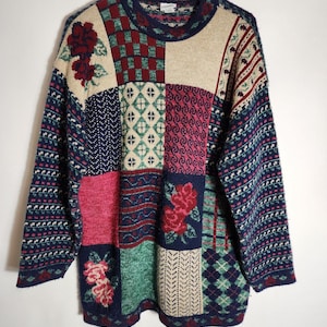 May include: A long-sleeve crew neck sweater featuring a multi-colored patchwork design. The knit pattern includes various blocks of cream, navy blue, dark red, and green, decorated with floral motifs, checkered patterns, and diamond shapes.