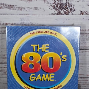 May include: A board game box titled "The 80's Game" with a blue background and a large yellow and red "80's" graphic. The text "How much do you remember?" is below the game title. The box is on a wooden surface.