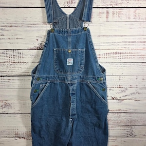 May include: Blue denim overall shorts with adjustable straps and metal hardware. Features a front bib pocket with a small patch and side pockets. The shorts are hanging on a white hanger against a light wood background.