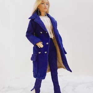 Op de afbeelding: Een pop gekleed in een koningsblauwe, getextureerde jas met bijpassende broek en witte laarzen. De jas heeft grote revers en zilveren knopen. De pop draagt ook een witte blouse en zilveren oorbellen. De outfit is een modieus ensemble.