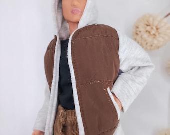 May include: A doll with a light brown complexion wearing a black shirt, brown corduroy pants, and a hooded jacket. The jacket features a light gray heathered jersey hood and sleeves paired with a dark brown quilted vest front panel.