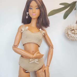 May include: A doll wearing a beige two-piece outfit with bow accents. The doll has brown hair, blue eyes, and is wearing earrings. The outfit includes a crop top and high-waisted bottoms. The background is a plain white wall with a few decorative leaves.