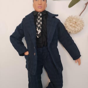 May include: A doll dressed in a navy blue suit, black shirt, and a black and white patterned tie. The suit has a textured finish. The doll has a neutral expression.
