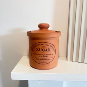 May include: A brown sugar canister with a lid. The canister has a circular label with the text "SUGAR" and "The Original Suffolk Canister." The label also includes "Ideal Storage at Home or on Safari" and "Henry Watson Pottery, England, Founded 1800."