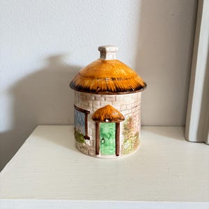 Peut inclure: Un pot en céramique en forme de chaumière avec un toit de chaume et une porte verte. Le pot présente un motif de briques beige clair et une petite fenêtre. Le toit est de couleur brun chaud, et le pot est posé sur une surface blanche.