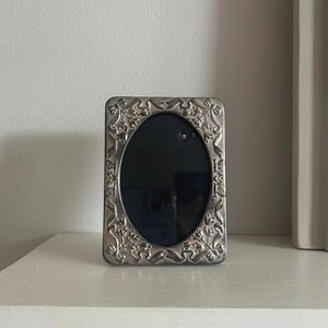 May include: A rectangular silver picture frame with an oval aperture. The frame has an elaborate floral and leaf pattern around the edges. The frame is displayed on a white surface.