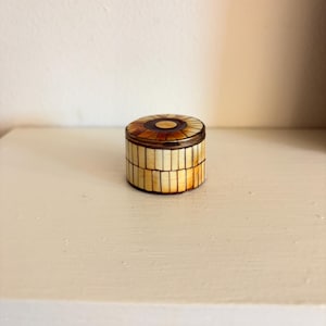 May include: A small, round decorative box with a lid. The box is made of light-coloured rectangular pieces, with a circular design on the lid featuring brown and tan hues. The box has a brass-coloured metal rim.