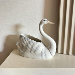 May include: A white ceramic swan planter with a long, curved neck and a slightly open beak. The swan's feathers are detailed with soft grey shading. The planter is sitting on a white surface.