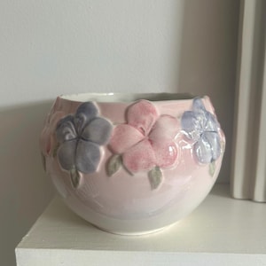 May include: A pale pink ceramic bowl featuring embossed floral patterns in pink, blue, and grey. The bowl has a rounded form and is placed on a white surface. The flowers are detailed, adding a decorative element.
